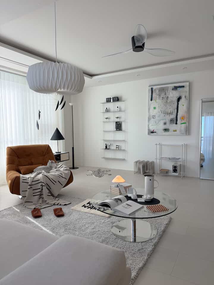 ホワイトとミニマルカラーのリビング、オレンジのアームチェアとグレーのラグが配置され、毛布の上で猫がくつろぐ雰囲気のある空間