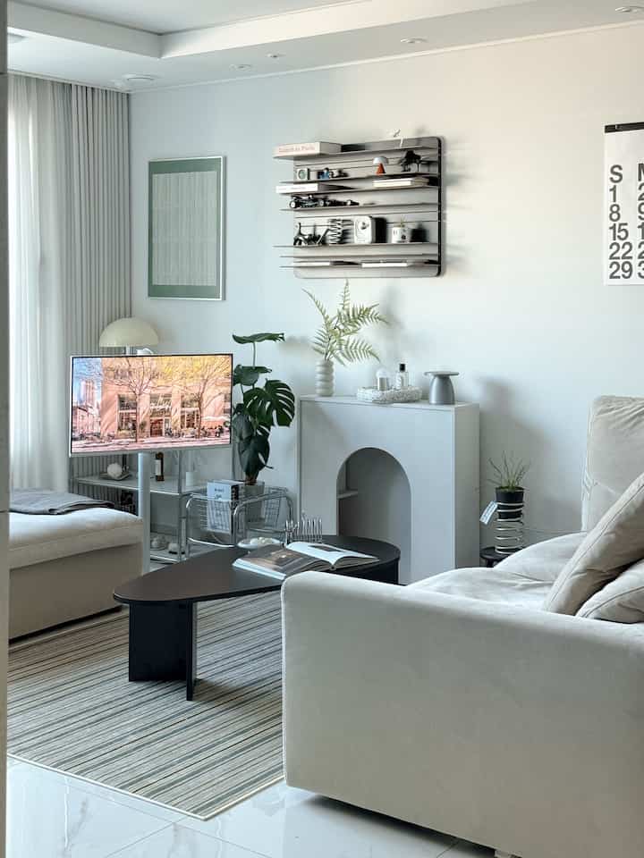 明るいホワイトトーンのリビング、ベージュのソファとブラックのコーヒーテーブル、中央にテレビ台と観葉植物が配置されたモダンで清潔感のあるインテリア
