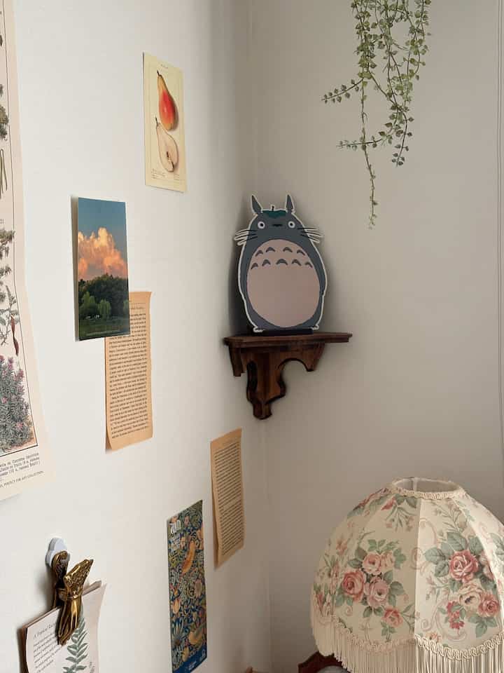 ナチュラルカラーの壁にフレンチカントリースタイルのウォールデコと小さなコーナー棚、ヴィンテージ調の花柄ランプのある雰囲気のある空間