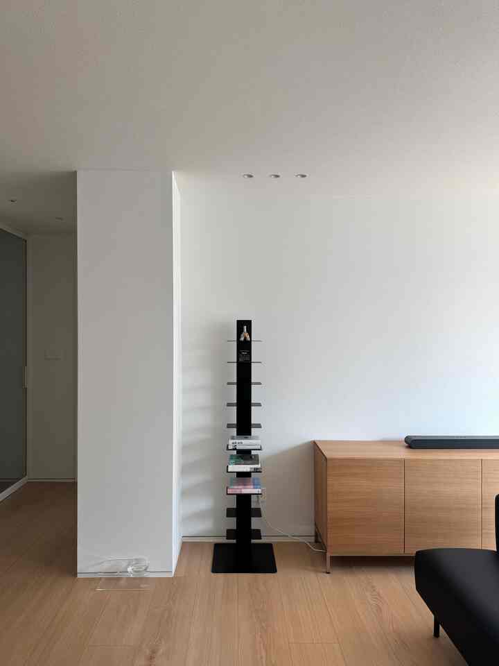 ナチュラルカラーでミニマルな印象のリビング、中央にスリムな黒い棚、右側に木製のテレビ台と黒いソファが配置された空間