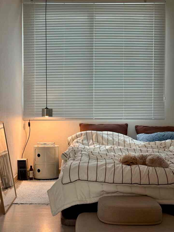 ホワイトとブラウンの色調の寝室、ブラインドが窓を覆い、ベッドに犬が横たわり、ナイトテーブルとペンダントライトが配置された落ち着いた空間