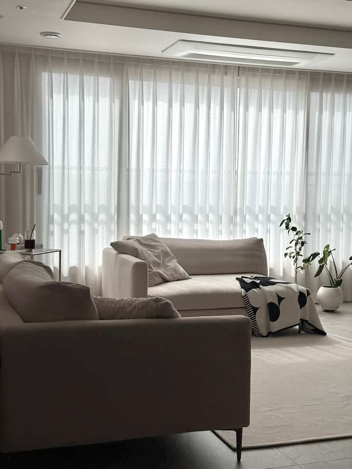 柔らかなベージュ色調のリビング、二つのソファと大きな窓にかけられたカーテンが配置された落ち着いた空間