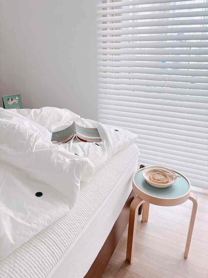 ホワイトトーンの寝室で、ボタンポイントの掛け布団とマットレスカバーが敷かれたベッドと木製スツールが配置された清潔感のある空間