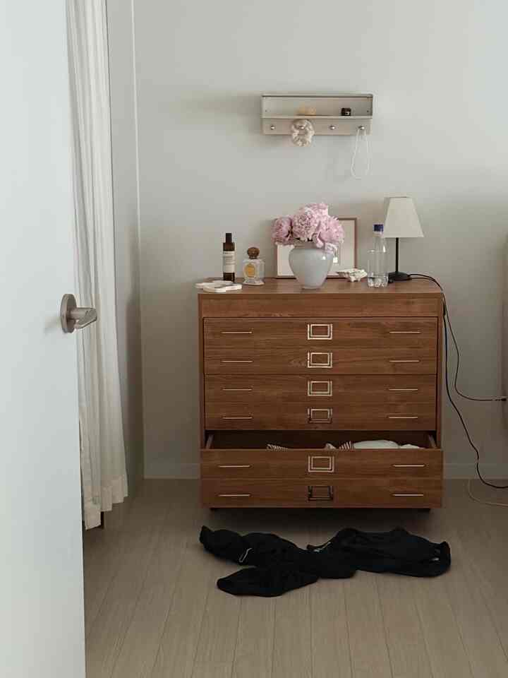 白い壁とナチュラルな木製チェストがある狭い部屋、花瓶とランプが配置された整った空間
