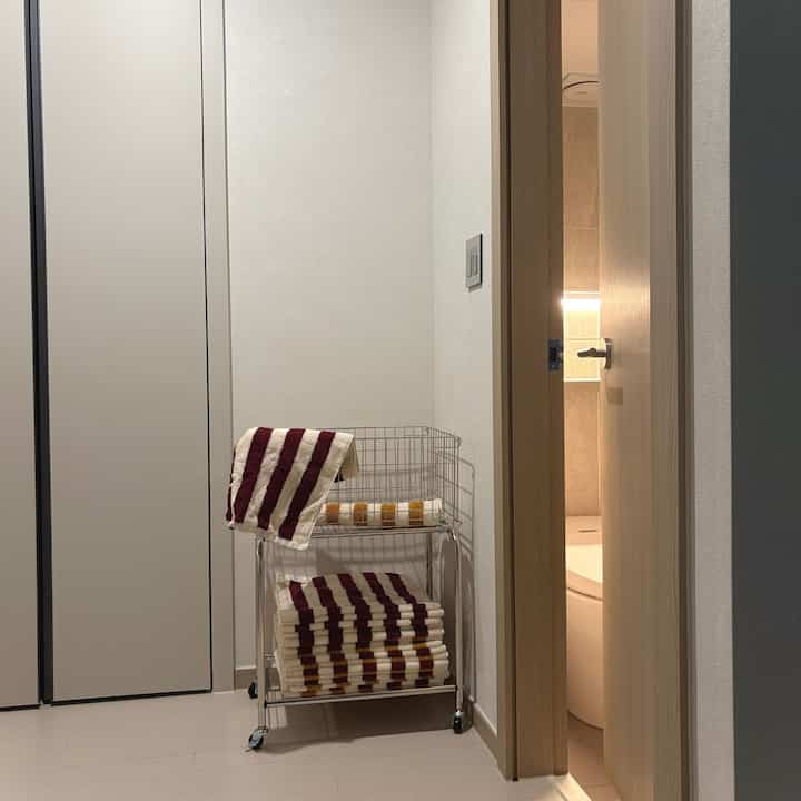 白色調のコンパクトな空間にタオルが置かれた移動式ワゴンと、開いた扉から見えるトイレがある整然とした収納スペース
