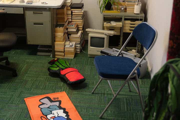 緑のカーペットが敷かれたコンパクトな書斎、青い折りたたみ椅子とレトロなコンピューターが配置された空間