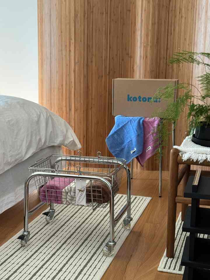 Cozy bedroom space featuring warm wood tone wall and flooring, white bed, stainless basket trolley cart with blue and pink towels