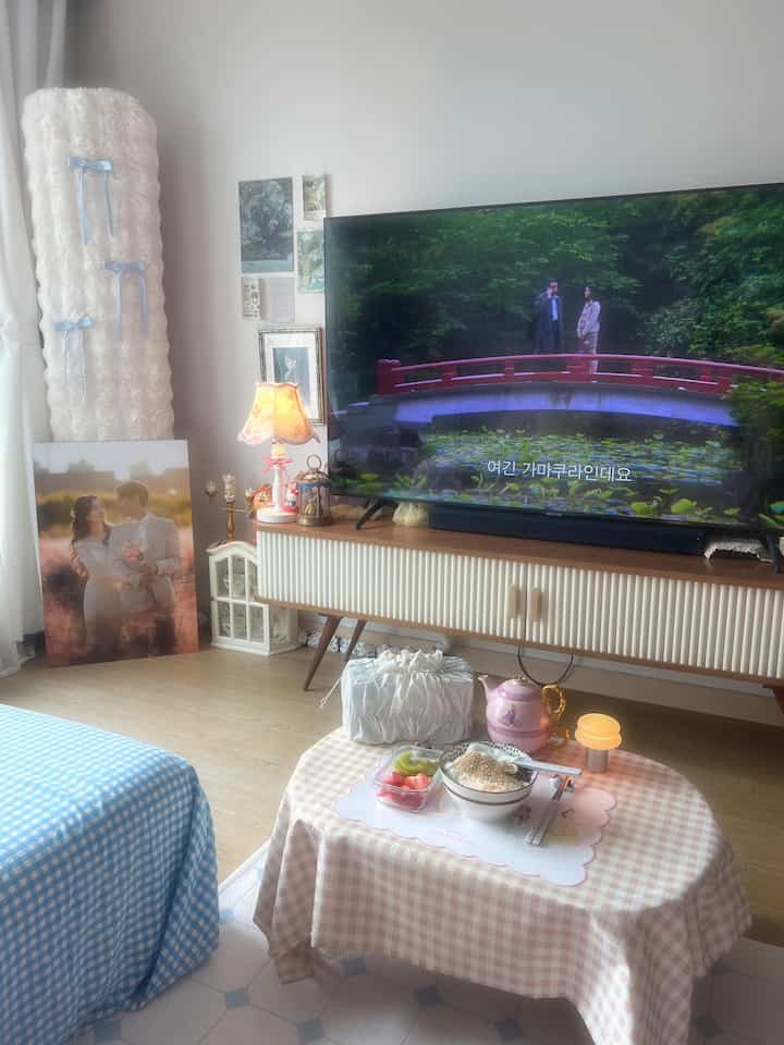 ベージュとブルーの色調のリビングルーム、チェック柄のベッドと額縁、テレビ、温かみのある照明が印象的な空間