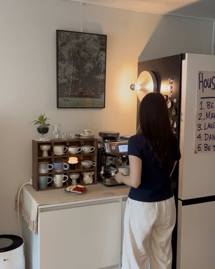 白と茶色のトーンのワンルームおうちカフェ、コーヒーマシンと整理されたマグカップの棚、壁の照明が配置された雰囲気のある空間