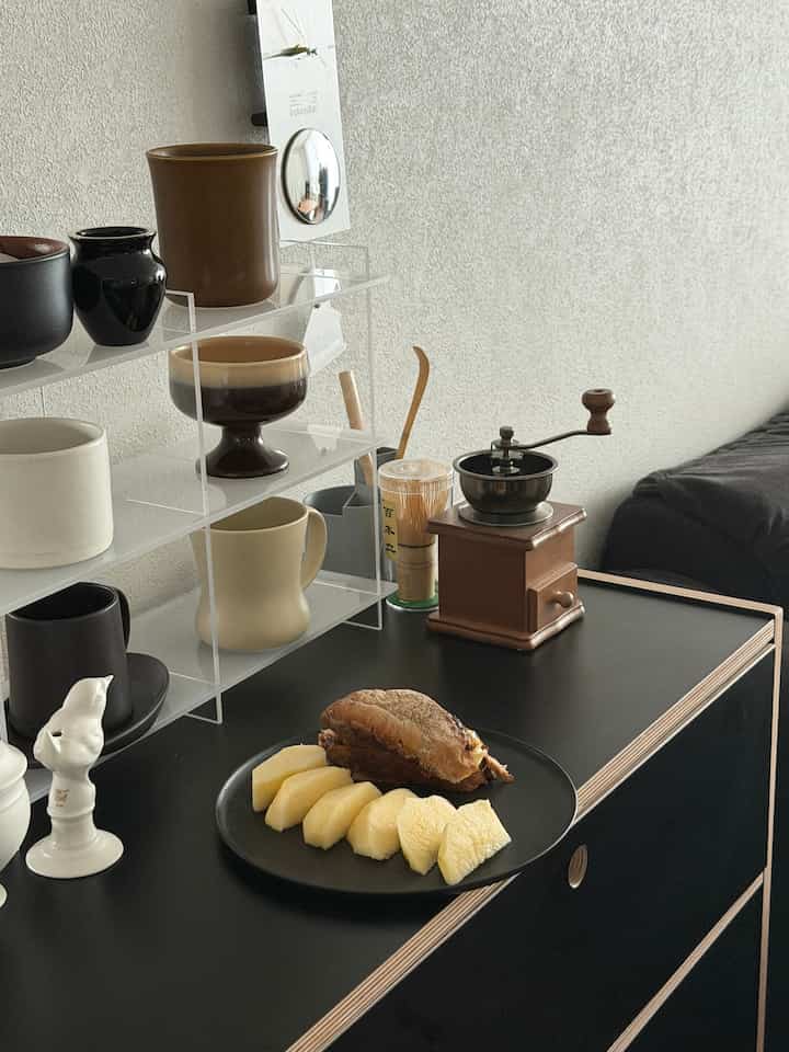 ナチュラルカラーのホームカフェ、様々なマグカップとコーヒーグラインダーが置かれたテーブルとアクリル棚が配置された雰囲気のあるインテリア