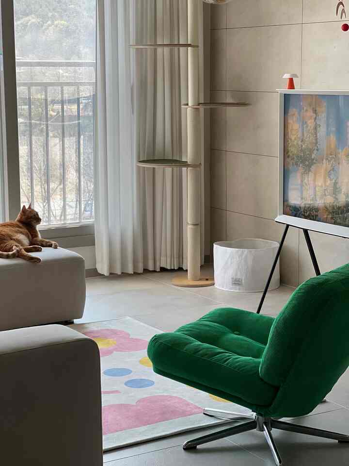 白いカーテンとベージュのキャットタワーがあるモダンなリビング、緑のアームチェアと花柄のラグが配置された雰囲気のある空間