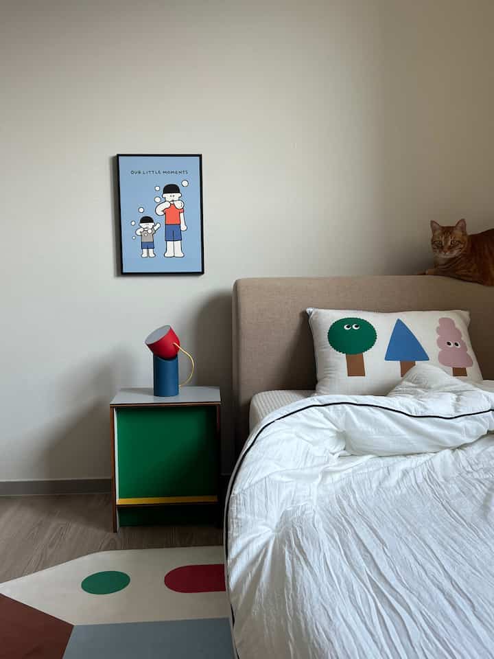 白い壁とベージュのベッドがある子供部屋、子供向けのポスターやカラフルなクッション、柄のラグ、ベッドのヘッドボードに猫がいる雰囲気のあるインテリア