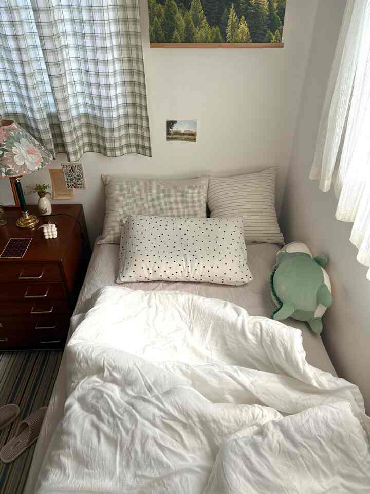 ナチュラルな色調の小さな寝室、白い寝具とナイトテーブル、チェック柄のカーテンが配置された温かみのある空間
