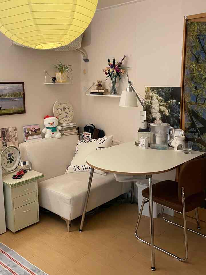 Bright beige floor and white furniture define a studio apartment home office and dining area, featuring a paper pendant light and wall shelves adding cozy atmosphere