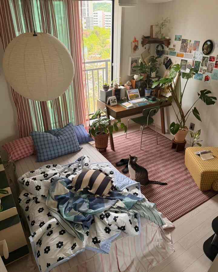 自然なグリーン植物と温かみのあるベージュのカーテンがある一人暮らし用寝室、木製のデスクと白い壁が居心地の良い雰囲気を演出している