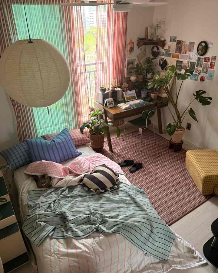 暖かいレトロでナチュラルな色調の寝室兼ホームオフィス、大きな窓辺にストライプのカーテンと木製デスク、多様な観葉植物と猫がいる居心地の良い空間