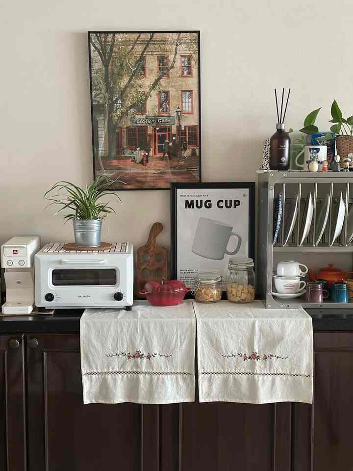 白と茶色のトーンのキッチンカウンター、トースターオーブンとコーヒーマシン、植物と額縁が配置された雰囲気のあるおうちカフェ空間