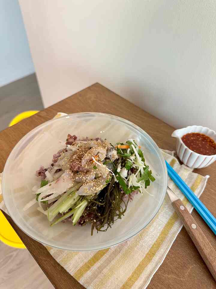 白い壁と木目調のダイニングテーブルがあるシンプルなワンルーム空間、中央に透明な器に盛られた新鮮な刺身丼が置かれている。