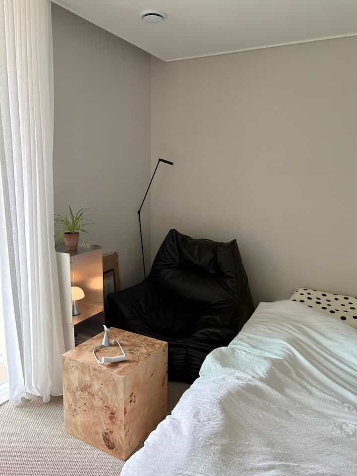 A cozy small bedroom with natural tones and beige walls featuring a black bean bag, bright curtains, and a wooden side table
