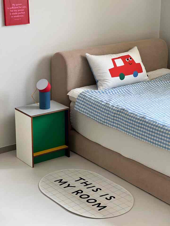 ベージュのベッドと青いチェック柄の布団がある子供部屋。かわいい赤い車の枕、白い壁、カラフルなスツールが配置された温かみのある空間。