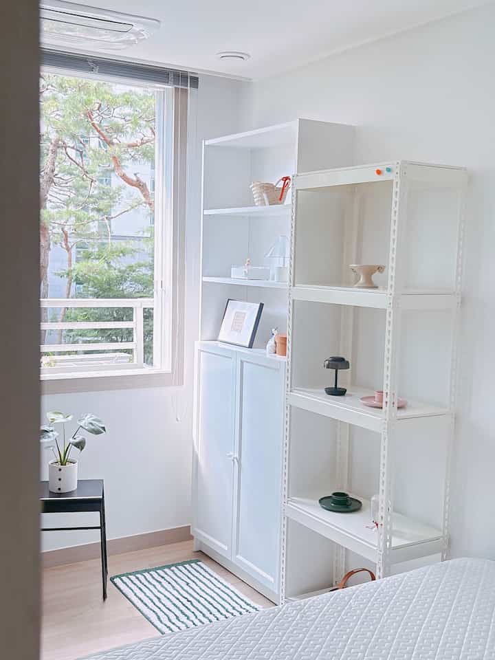 明るい白とアイボリーの色調が特徴の小さな寝室、金属製の棚とビリー本棚が配置されたミニマルで清潔感のあるインテリア