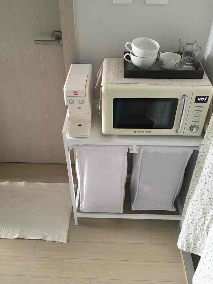 ホワイトとナチュラルのトーンでまとめられた狭いキッチンスペース、電子レンジとコーヒーマシンが置かれた棚と整頓されたランドリーバスケットが配置された実用的な空間