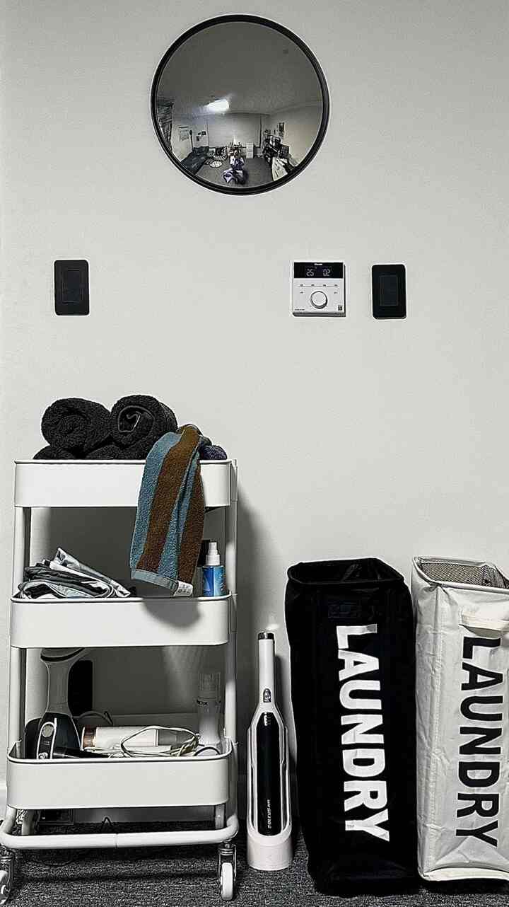 ホワイトの壁にブラックのスイッチと丸い鏡、ホワイトの収納トロリーとブラック＆ホワイトのランドリーバスケットが置かれたモダンなバスルーム空間