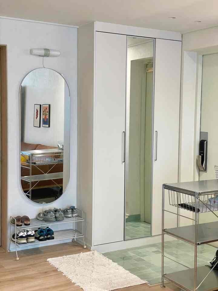 ホワイトとライトグレー調の玄関、広いシューズラックとメタル棚、縦長の楕円形ミラーが機能的な収納と清潔感ある雰囲気を演出するインテリア。