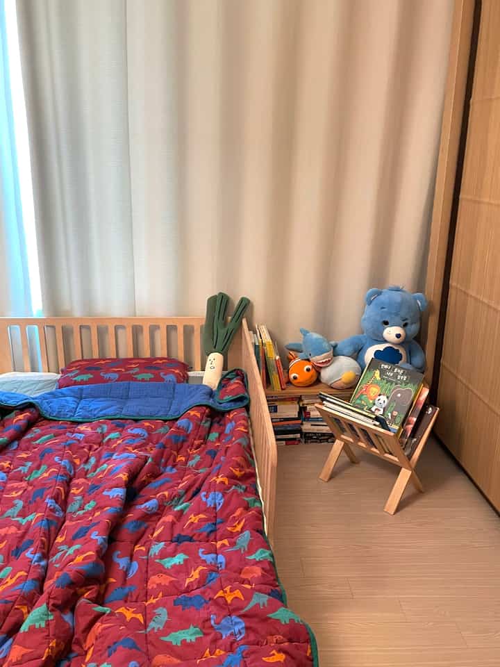 ナチュラルな色調の子供部屋、木製ベッドにカラフルな恐竜柄の寝具、ぬいぐるみと棚が配置された雰囲気のあるインテリア