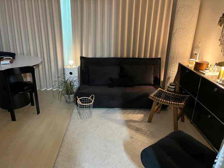 ベージュのカーテンを背景にブラックのソファと木製スツール、白いラグが配置された雰囲気のあるリビング