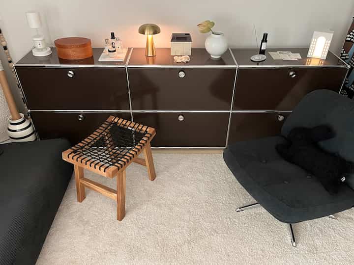 ナチュラルトーンのリビングスペース、壁沿いに配置されたUSMキャビネットと木製スツール、ダークグレーのアームチェアが調和したモダンで清潔感のあるインテリア