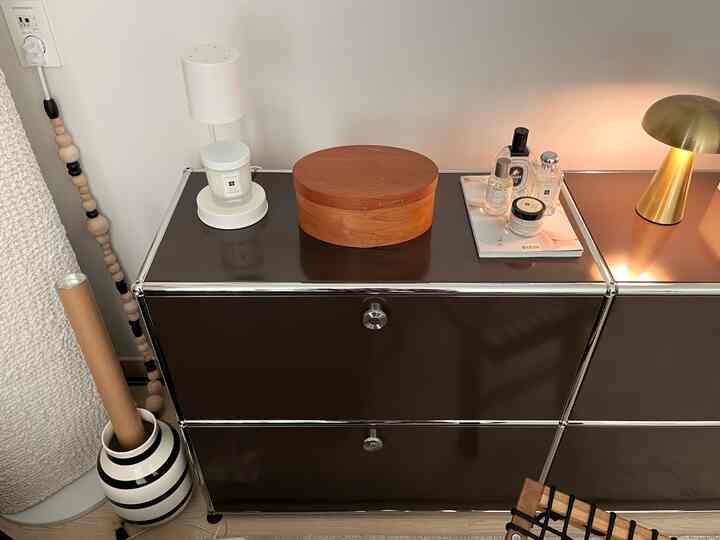 ブラックとシルバーのUSM収納の上に木製のシェイカーボックスと香水、ゴールドのテーブルランプが配置されたシンプルでモダンなオブジェ空間