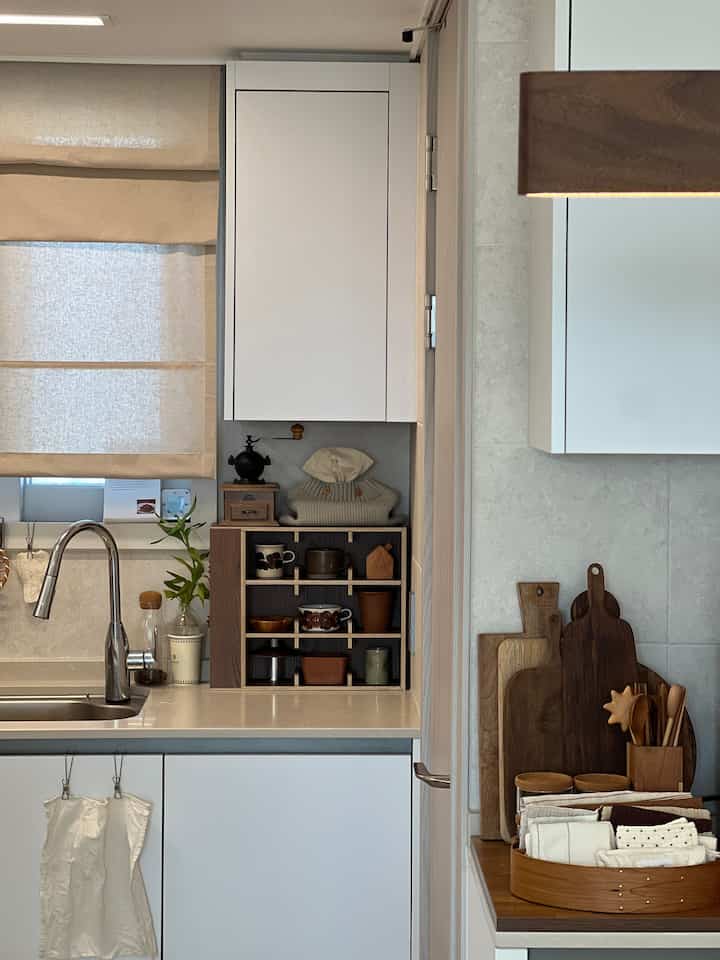 白と茶色のトーンのキッチン空間、シンクと木製のカップ棚や調理器具が配置された自然で清潔感のあるインテリア
