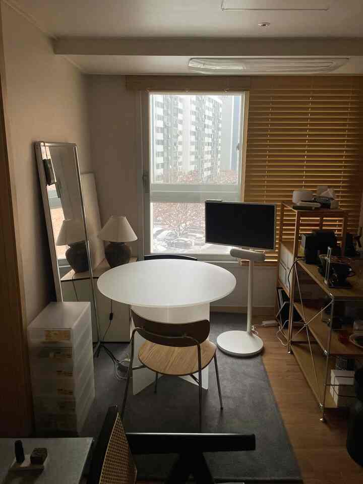 ウッドトーンとホワイトが調和したコンパクトなリビング、丸いテーブルと椅子、ウッドブラインドが配置されて温かみのあるインテリア