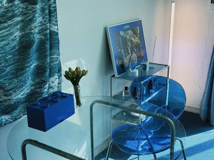 ブルーの透明な椅子とガラステーブルが配置されたミッドセンチュリーモダンスタイルの小さなリビング空間、ヴィンテージのパリポスターが飾られたクールな印象のインテリア