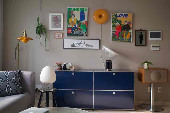 ナチュラルカラーの壁とモダンな家具が配置されたリビング、グレーのソファとネイビーのキャビネット、ポスターと額縁付きアートが調和したシンプルな空間