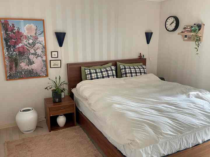 ホワイトとブラウンの色調の寝室、キングサイズベッドとナイトテーブル、壁付けライト、時計が配置された落ち着いたナチュラルな空間