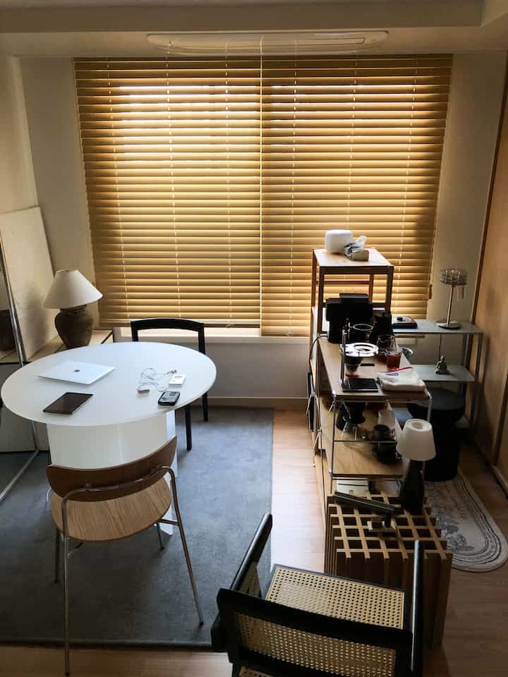 ナチュラルなウッドトーンと温かみのあるブラインドが印象的なコンパクトなホームカフェ空間、丸いダイニングテーブルとステンレス棚が配置されたモダンなインテリア