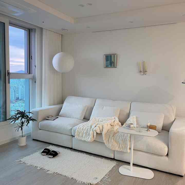 白を基調としたリビング、広いモジュールソファとラグ、カーテン、サイドテーブルが配置されたモダンで清潔感のあるインテリア