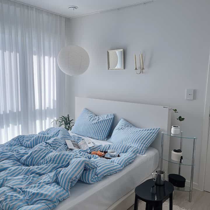 白と青のトーンの寝室、すっきりとしたベッドとカーテン、コーナー棚と植物が配置されたシンプルな空間