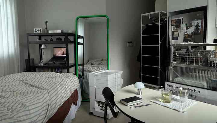 モノトーンのコンパクトな寝室、緑のフレームの鏡とテーブルランプ、デスクが配置されたモダンなインテリア