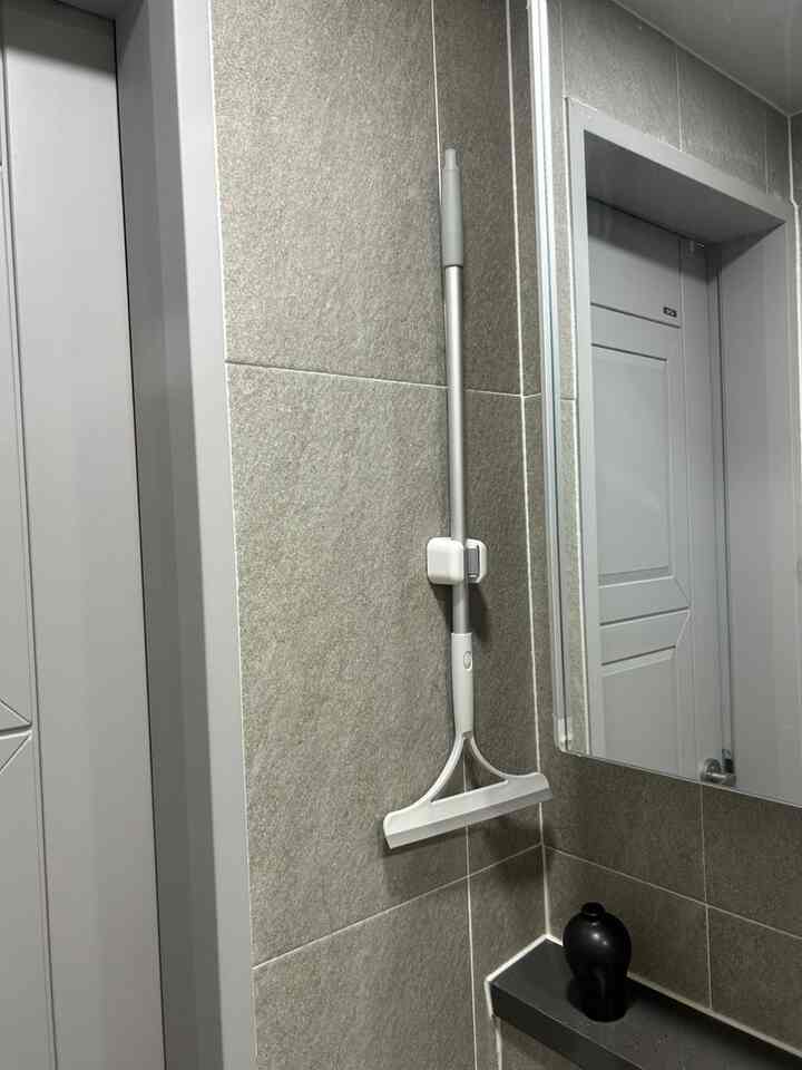 グレーのタイル壁に取り付けられた白いスクイージーとモップホルダーが見える、清潔な縦長のトイレのコーナー空間