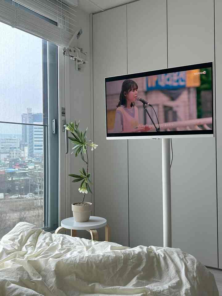 白とナチュラルカラーの寝室、窓辺に置かれた木製スツールの上の植木と白いベッドリネンが落ち着いた雰囲気のインテリア