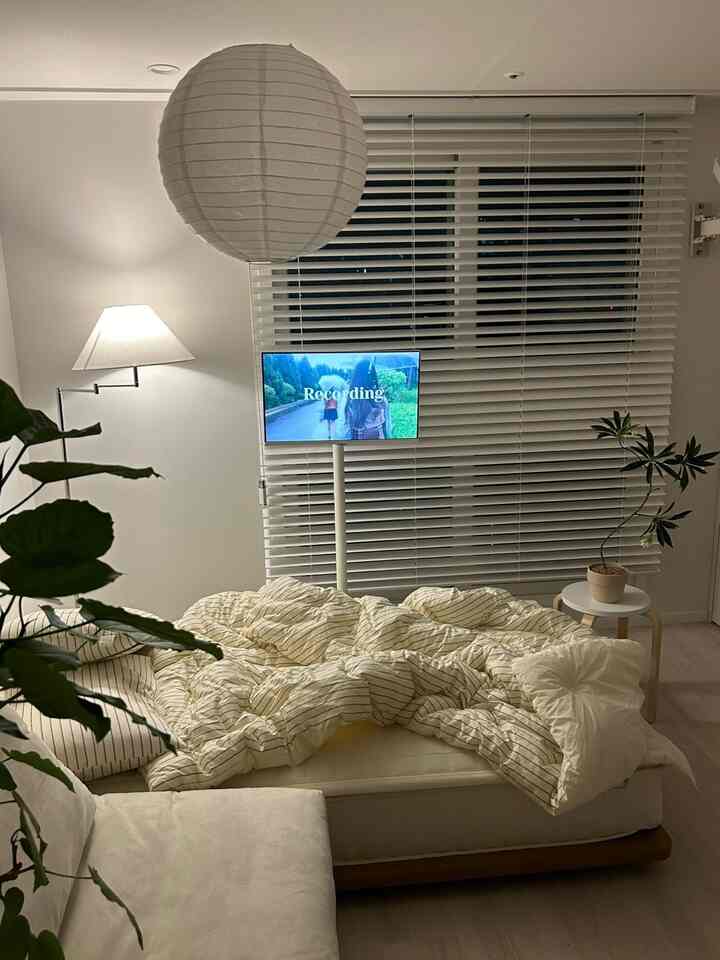 やわらかな白とアイボリーの色調の寝室、低めのベッドフレームと木製ブラインド、フロアランプで落ち着いた夜の雰囲気のあるインテリア