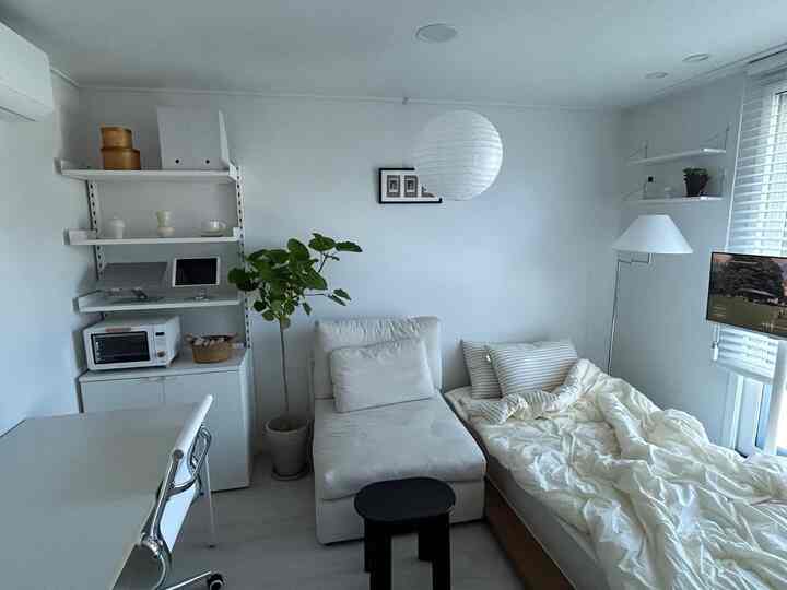 ホワイトとベージュのトーンでまとめられたコンパクトな寝室、棚や植物、フロアランプが配置されたミニマルなインテリア