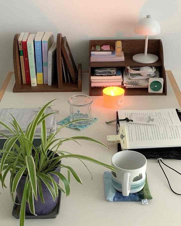 ホワイトのテーブルクロスが敷かれたデスクに、本、ランプ、キャンドル、植物、マグカップが配置された雰囲気のある読書空間
