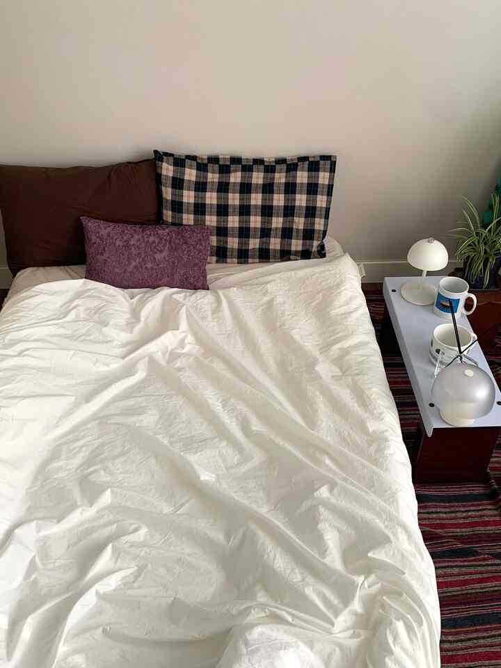 白い寝具と茶色のクッションが配置された寝室、カスタムミニベンチのサイドテーブルと植物がある雰囲気のあるインテリア