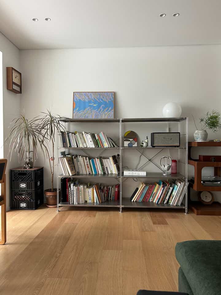 ナチュラルな色調のリビング、金属製の本棚に多様な本や植物、時計、ソファが配置された雰囲気のあるインテリア