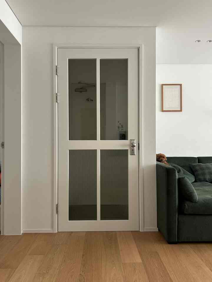 白い壁と木目調の床があるリビング、透明およびすりガラスの中間ドア、ダークグリーンのソファに小型犬が座っている雰囲気のあるインテリア