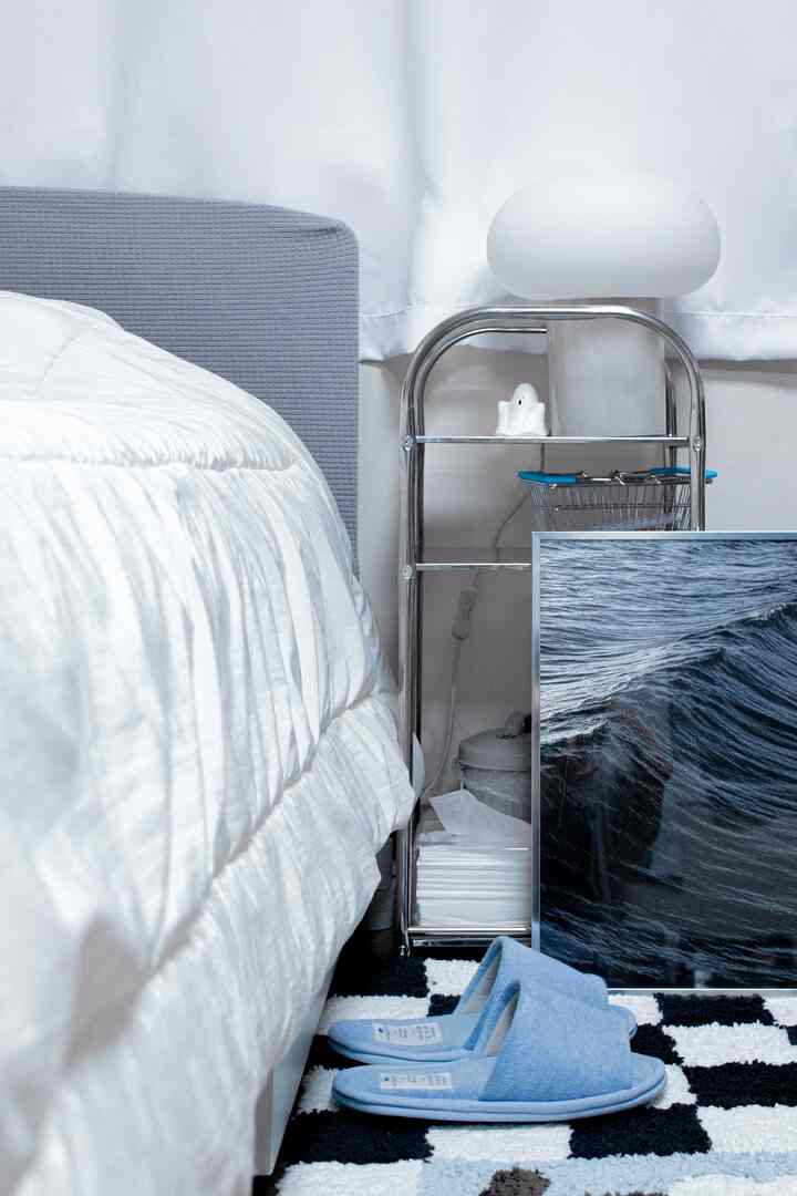 白と青のトーンの寝室、チェック模様のバスマットとステンレス製のサイドテーブル、波のポスターが配置された落ち着いた雰囲気のインテリア